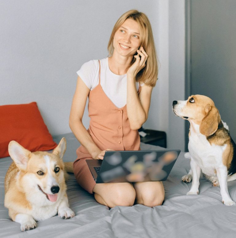 Smiling woman working on laptop with dogs at home, enjoying remote work on bed.